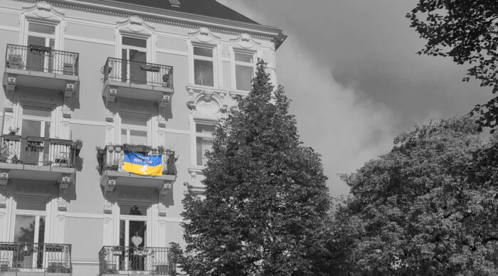 Foto eines mehrschössigen Eckhauses, daneben Bäume und der Himmel. Das Bild ist in schwarz-weiß, außer einer gelb-blauen Ukraine-Flagge, die in der Mitte des Bildes von einem Balkon hängt.