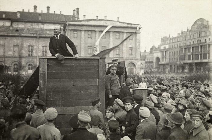 Schwarzweißfoto von Lenin, der auf einem Holzpodest stehend auf einem Platz eine Rede hält, Hunderte Männer, viele in Uniform, hören zu