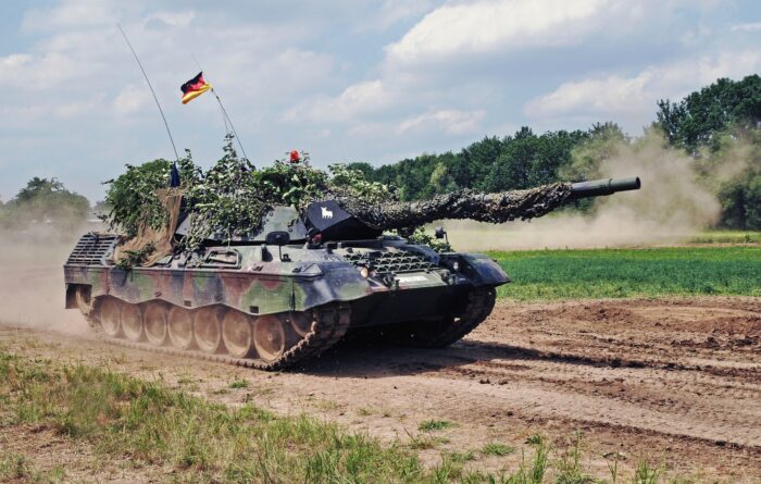 Ein Panzer mit Tarnnetz, Ästen und Deutschlandfahne fährt über eintrockenes Feld