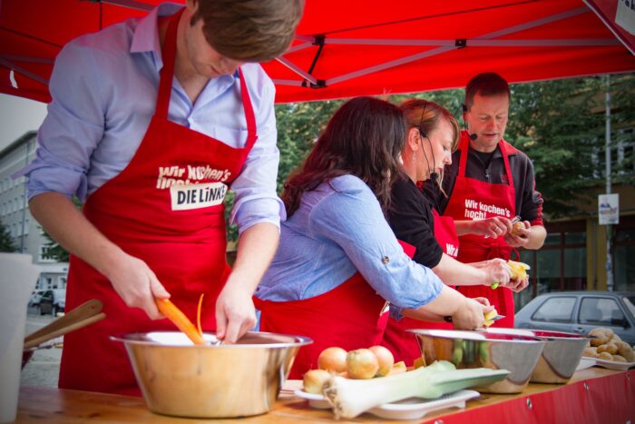 Mehrere Menschen mit roten Schürzen mit der Aufschrift "Wir kochen's hoch. Die Linke" schneiden Gemüse an einem Stand