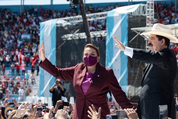 Eine Frau mit Corona-Maske steht vor einer Menschenmenge und winkt, rechts neben ihr ein Mann mit Schnauzer und Cowboyhut, der ebenfalls winkt, im Hintergrund die Flagge von Honduras