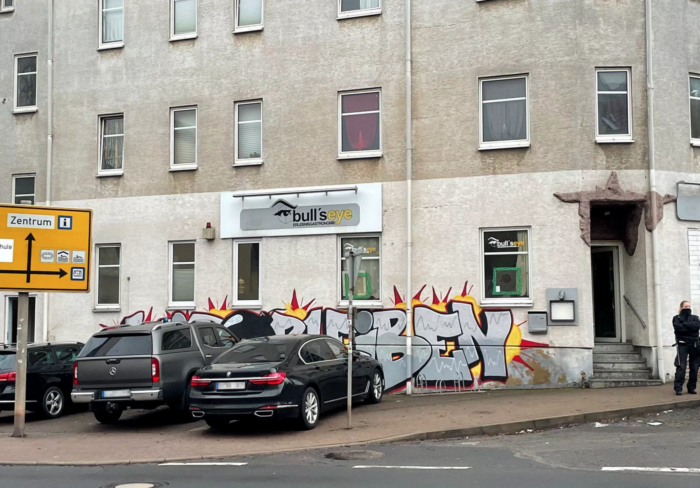 Ein graues Haus mit Eingang zu einer Kneipe. An der Wand ist ein Schild "Bull's Eye" angeschraubt, vor der Kneippe stehen einige dunkle Autos, vor der Tür eine Polizistin.