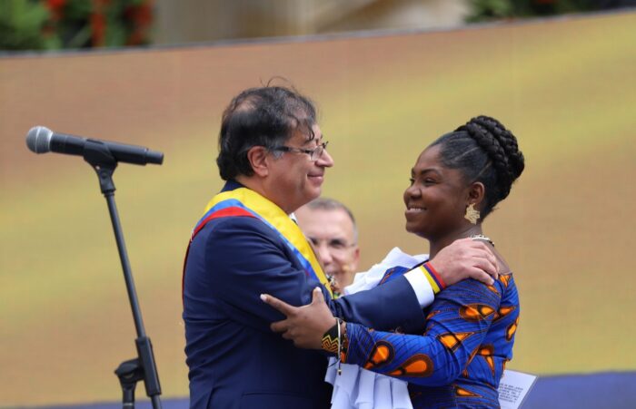 Ein mittelalter Mann mit einer Schärpe in den Farben der kolumbianischen Flagge und eine jüngere Schwarze Frau umarmen sich, links von ihnen steht ein Mikrofon