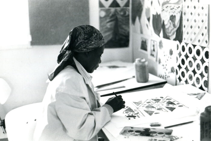 Bild einer Frau im weißen Mantel und Kopftuch, die gerade geometrische Zeichnungen auf einem Blatt Papier macht. Sie ist umgeben von Skizzen, die an die Wände hängen.
