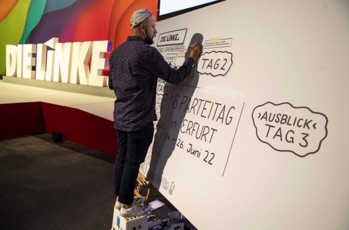 Ein Mann steht auf einem hocker und zeichnet etwas auf eine Papierwand, man kann die Worte "Parteitag" und "Ausblick Tag 3" erkennen. Im Hintergrund ein großer Aufsteller des DIE LINKE Schriftzugs