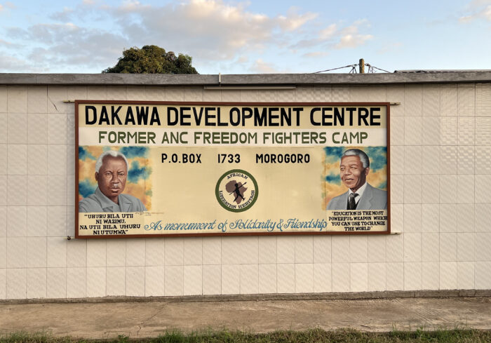 Bild eines an einer Wandmauer angebrachten Poster des "Dakawa Development center". Sarauf zu sehen ist in der Mitte das Logo der Bildungstätte. Links ein gezeichnetes Porträt von Julius Nyerere, rechts eins von Nelson Mandela