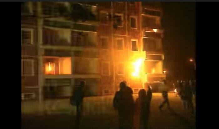 Nachtaufnahme: Flammen an einem Gebäude, davor schemenhaften Gestalten
