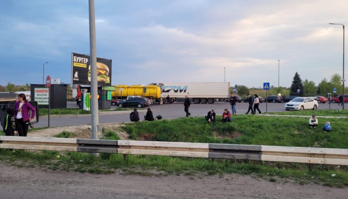 Ein Tankstellenparkplatz an der Autobahn, auf der Wiese davor sitzen zahlreiche Menschen.