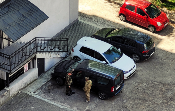 Ein Parkplatz, von oben aufgenommen. Neben einem der geparkten Autos stehen Männer in Uniform.