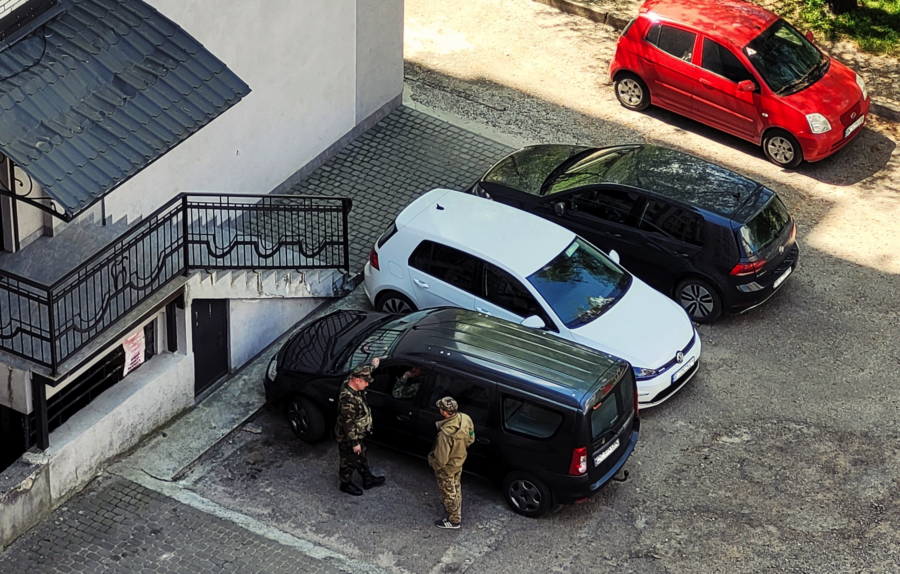 Ein Parkplatz, von oben aufgenommen. Neben einem der geparkten Autos stehen Männer in Uniform.
