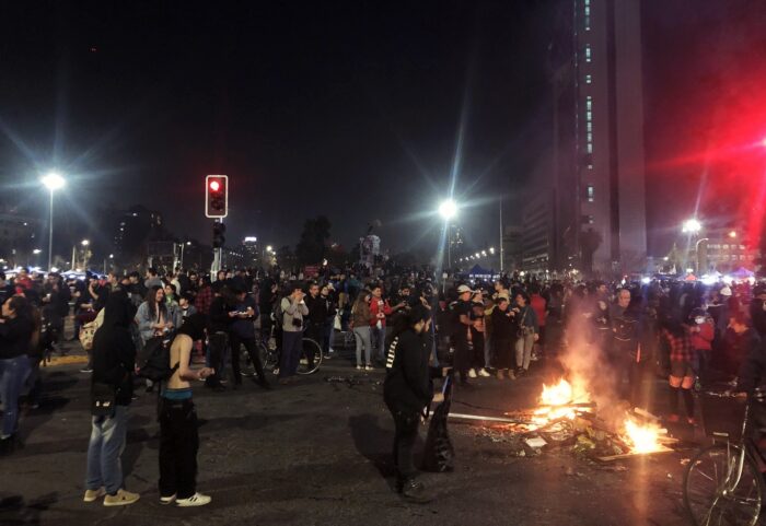 Eine zerstreute Menschenmenge in der Nacht auf einen Platz, am rechten Bildrand brennt ein kleines Feuer