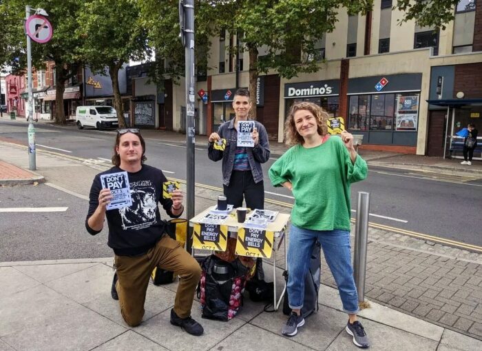 Drei junge Menschen halten an einem Stand an einer Straße Flyer hoch mit dem Slogan "Don't Pay" und "Don't Pay Energy Bills, Oct. 1"