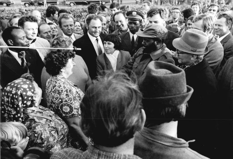 Foto einer Menschenmenge, in der Mitte unterhalten sich vier Leute. Links zweei Bäuerinnen und rechts Samora Machel in Militäruniform und daneben Zk-Präsident Erich Honeker.