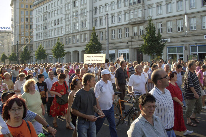 Eine Menschenmenge zieht durch eine Straße, auf einem Schild steht "1989: Wahlbetrug, 2004: Wahlbetrug"