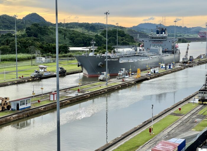 Ein großes (leeres) Containerschiff in einer Anlegestelle in einem Kanal, im Hintergrund grüne Hügel und ein weiteres, mit Containern beladenes Schiff