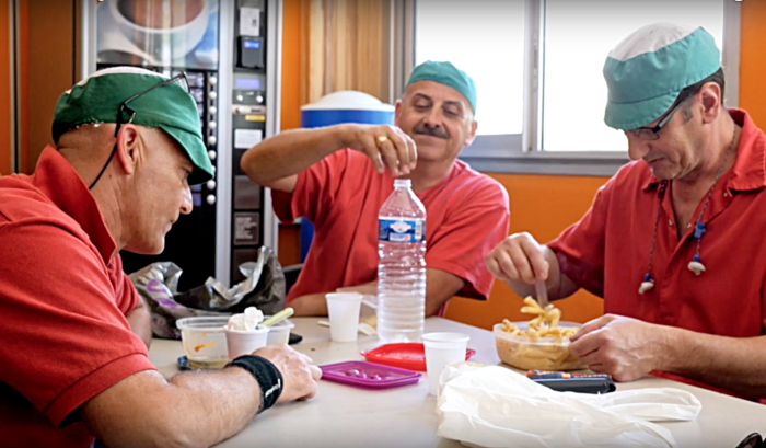 Drei Männer in Arbeitsuniformen sitzten um einen Tisch, einer isst aus einer Tupperdose Nudelsalat, einer schraubt eine Falsche zu