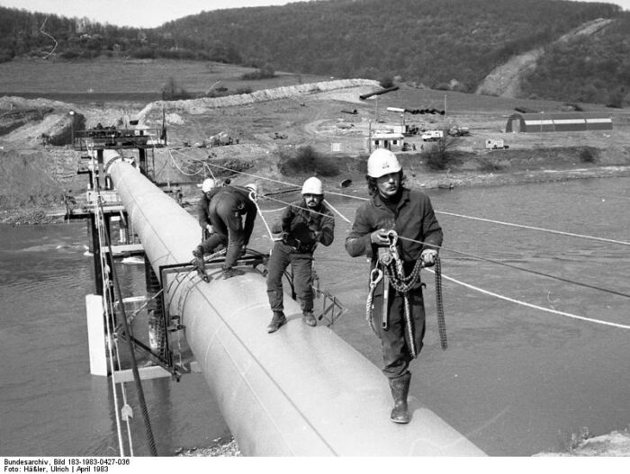 Drei Arbeiter beim Pipelinebau über einen Fluss.