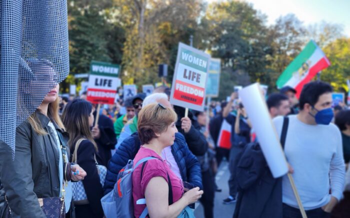 Demoszene, im Vordergrund links eine Frau, deren Gesicht von einer Fahne halb verdeckt ist, in der Mitte eine ältere Frau im Profil, rechs ein Mann mit Mundschutz, mehrere Fahnen und Schilder sind zu ehen, Aufschrift auf den Schildern: Woman, Life, Freedom