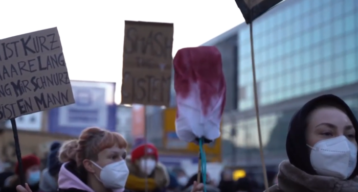 Mehrere Personen mit Masken halten im Rahmen einer Demonstration Schilder und einen gebastelten, rot gefärbten Riesentampon in die Höhe.