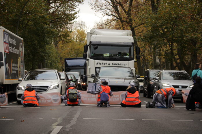 Einige Aktivist*innen der Letzten Generationen blockieren die Puschkin-Allee in Berlin. Vor ihnen ein weißer LKW