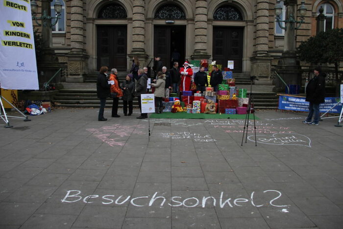 Mahnwache vor einem Gebäude. Mittig ist ein Berg Geschenke und circa 20 Leute, eine Person ist als Weihnachtsmann verkleidet. Eine andere Person filmt das Geschehen. Im Vordergrund ist "Besuchsonkel?" mit Kreide auf den Fußweg geschrieben.