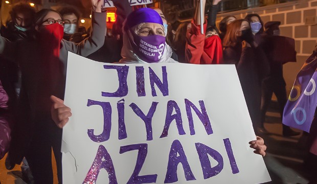 Man sieht eine Person mit Maske und einem großen Schild, auf dem steht Jin Jiyan Azadi.