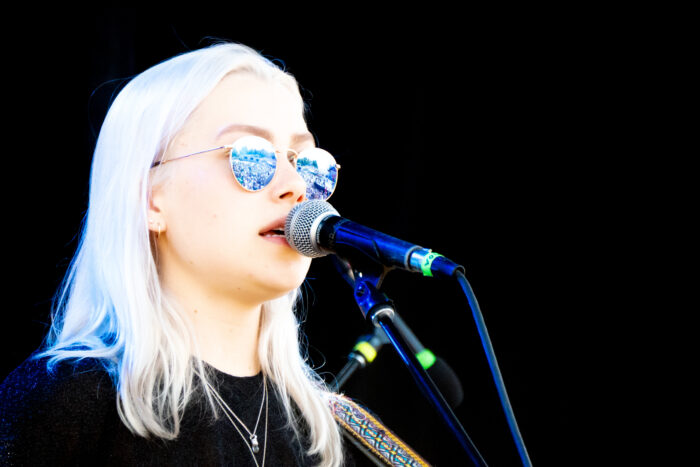 Eine junge Frau mit weißen Haaren und verspiegelter Sonnenbrille steht an einem Mikrofon und scheint zu singen.