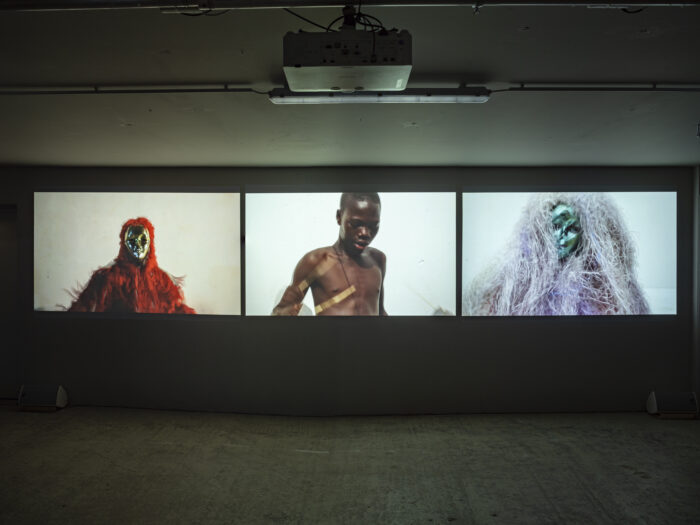 Bild einer Installation bestehend aus drei Bildschirmen. Rechts ein Tänzer mit Maske und rotem stoff. In der Mitte ein Schlagzeugspieler, rechts ein anderer Tänzer mit Maske