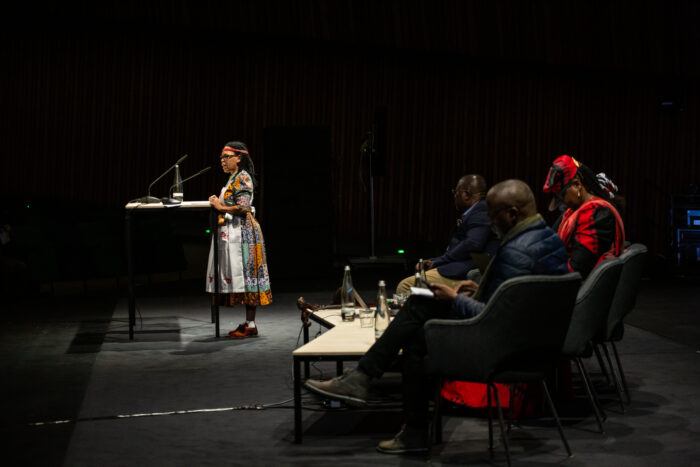 Eine Sprecherin steht auf einer Bühne am Pult und spricht über Mikrofone an ein Publikum, das nicht auf dem Foto ist. Sie trägt ein buntes Kleid. Rechts neben ihr sitzen weitere Sprecher*innen.