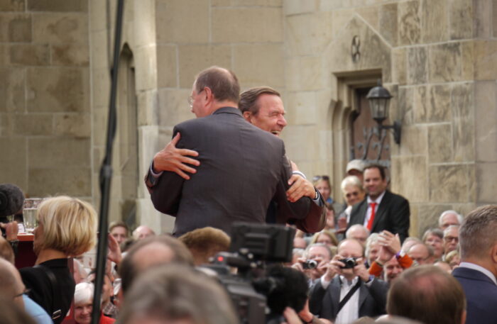 Peer Steinbrück und Gerhard Schröder umarmen sich auf einer Bühne, umgeben von Publikum. Schröder lacht.