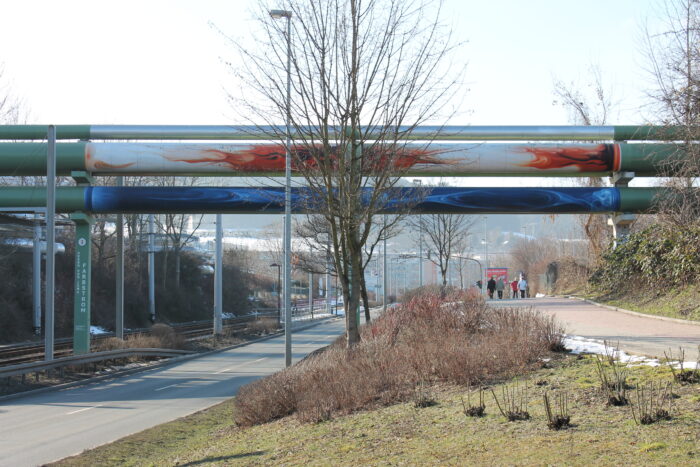 Ein künstlerisch verziertes Rohr einer Fernwärmeleitung, das über eine Straße verläuft.