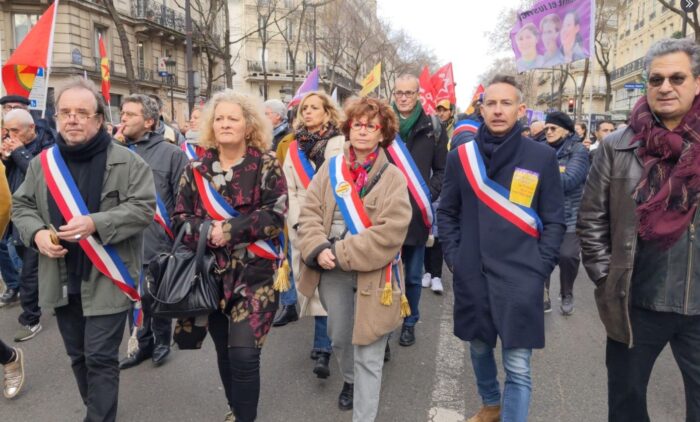 Man sieht fünf Personen, die nebeneinander auf einer Demonstration mit laufen. Vier von ihnen tragen eine Binde in den französischen Farben.