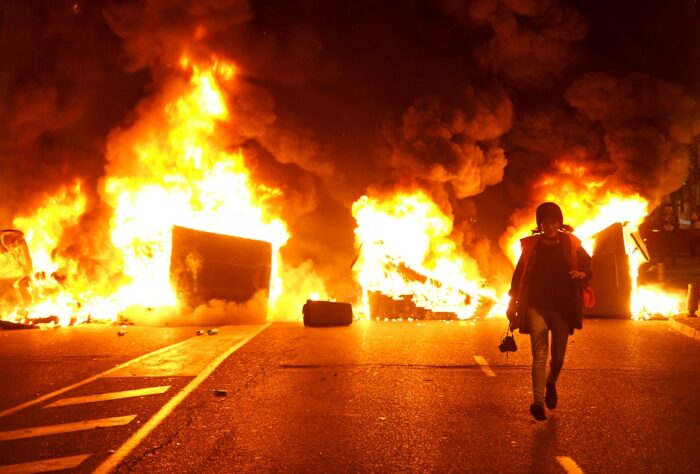 Brennende Mülltonnen, lodernde Flammen, dunkler Rauch. Von dieser Szene kommt eine Frau mit Fotoapparat den Betrachter*innen entgegegen.