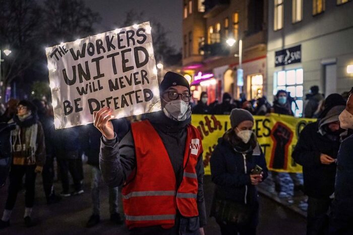 Es ist dunkel. Man sieht eine Person mit einem glitzernden Schild, auf dem steht: The workers united will never be defeated.