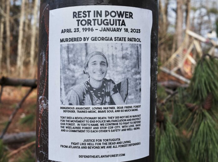 Auf einem Mast klebt ein Plakat auf dem ein Schwarzweißbild einer lächelnden Person zu sehen ist. Aufschrift: "Rest in Power Tortuguita. April 23, ,1996 - January 18, 2023. Murdered by Georgia State Patrol." Kleiner steht unter dem Bild: Indigenous anarchist, loving partner, dear friend, forest defender, trained medic, brave soul and so much more. Tort died a revolutionary death, They did not die in vain but for the movement to end police militarization and protect our forest. In Tort's name, we continue to fight to defend the Welaunee Forest and stop Cop City. With love, rage and commitment to each other's safety and well-being. Justice for Tortuguita. Fight like hell for the dead and living, From Atlanta and beyond. we are all forest defenders. defendatlantaforest.com