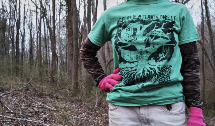 Eine Person mit grünem Shirt und pinken Arbeitshandschuhen stemmt eine Hand in die Hüfte, der Kopf ist nicht zu sehen, im Hintergrund Wald