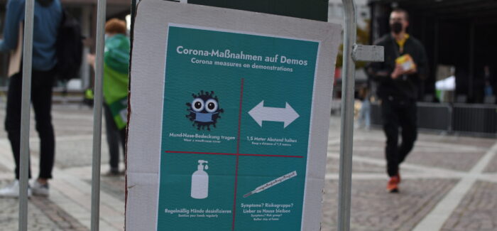 Plakat für Corona-Schutzmaßnahmen auf Demos. Ein stilisiertes Virus mit einer Maske und dem Text "Mund-Nase-Bedeckung tragen". Ein Pfeil, das in zwei Richtungen zeigt und dem Text "1,5 Meter Abstand halten". Ein Symbol einer Desinfektionsflasche und dem Text "regelmäßig Hände desinfizieren" und einem Thermometer mit dem Text "Symptome? Risikogruppe? Lieber Zuhause bleiben"