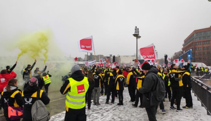 Man sieht eine Gruppe von Menschen von hinten, mit gelben verdi Westen und Fahnen, sie stehen im Schnee, die Luft ist nebelig.