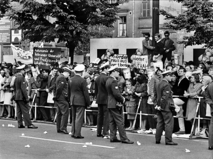 Foto einer Demonstration. Auf der Straße stehen Polizisten vor einer Barriere. Hinter der Barriere stehen dicht einander Demonstrantinnen und Demonstraten.