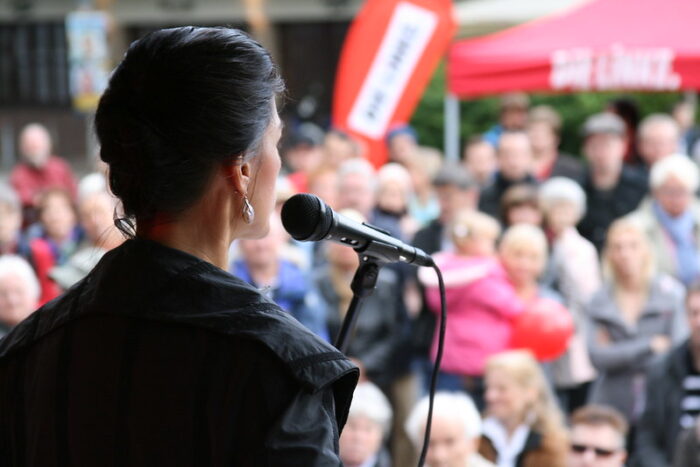 Auschnitt aus einer Demo, von der Bühne. Im Vordergrund spricht Sarah Wagengnecht in ein Mikrophon zu einer Gruppe vor ihr, im Hintergrund ein Stand der Linkspartei.