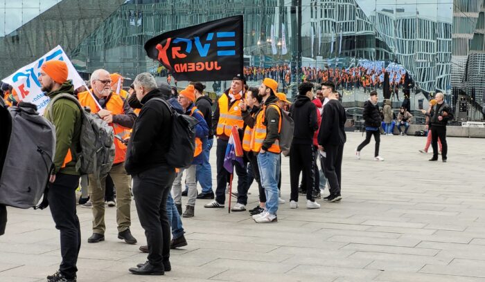 mehrere Leute in orangefarbenen Streikwesten stehen auf einem PLatz, eine Gruppe hält ein Schild mit der Aufschrift EVG-Jugend, im Hintergrund spiegelt sich die Streikkundgebung in einer Glasfassade