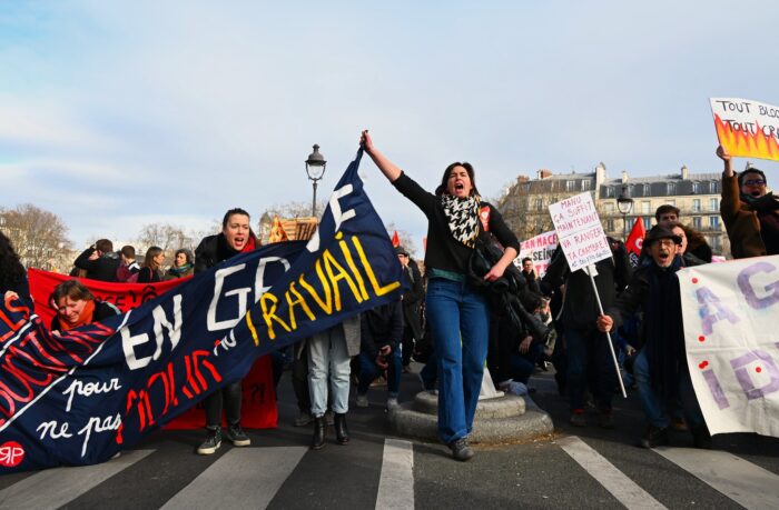 Eine Demonstration, von vorne, man sieht eine FRau ein transparent hochhalten und etwas rufen, drumherum mehr Leute mit Schildern, die ebenfalls rufen