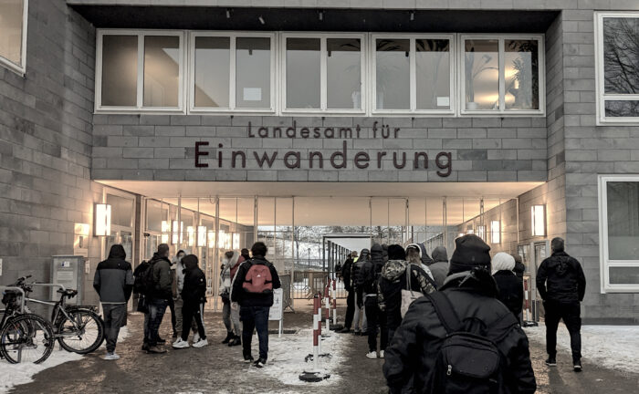 Frontalansicht eines Gebäudeeingangs im Winter. Eine lange Schlange wartet im Schnee vor einem Gebäude, auf fdem steht "Landesamt für Einwanderung"