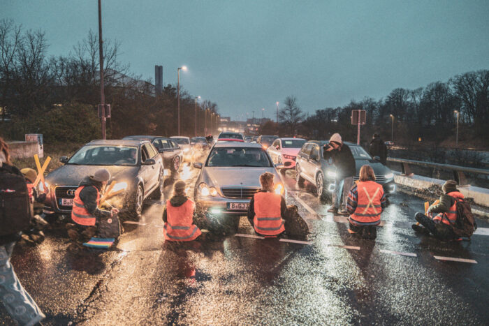 Vier Mitglieder der Letzten Generation mit roten Warnwesten sitzen auf einer Straße und blockieren viele Autos.