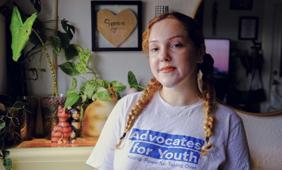 Eine junge Frau mit zwei Zöpfen steht in einer Wohnung, uf ihrem Shirt steht "Advocates for Youth", im Hintergrund ein paar Pflanzen und Katzenfiguren