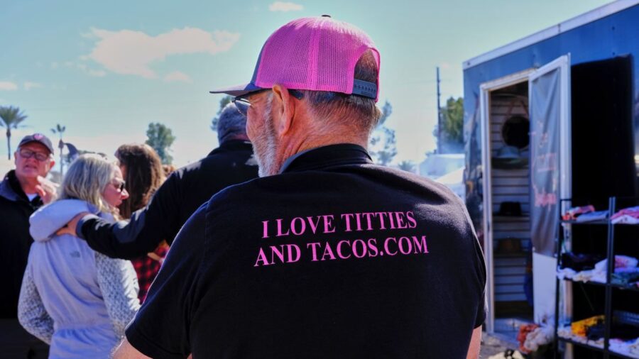 Ein Mann steht mittig mit dem Rücken zur Kamera. Er trägt ein pinkes Cap. Auf seinem T-Shirt steht in pinker Schrift I love titties and tacos . com