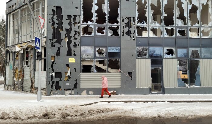 Eine Person in rosaroter Winterkleidung geht eine verschneite Straße entlang, vorbei an einer Fassade, an der sämtliche Fenster zerstört sind