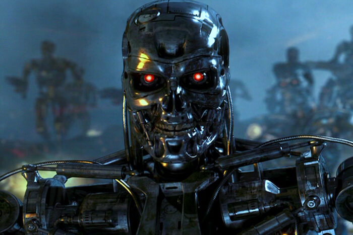 Bildausschnitt aus dem Terminator. In der Mitte des Bildes eine Nahaufnahme eines Roboters aus glänzendem Metall und einem Kopf, der aussieht wie ein menschlicher Skelett und roten Augen.