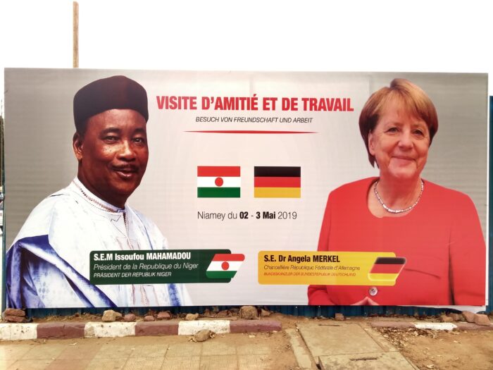 Bild eines überdimensionierten Schildes am Straßenrand. Darauf zu sehen sind rechts Angela Merken und links Nigers Präsident Issoufou Mahamadou, in der Mitte die deutsche und nigrische Flagge, dazum 02 bis 03 Mai. Betitelt ist die Veranstaltung auf deutsch und Französisch als "Besuch von Freundschaft und Arbeit"