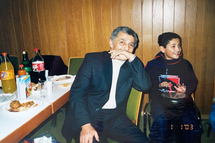 Ein älterer Mann in Anzug sitzt an einem Tisch, neben ihm ein kleiner Junge, beide schauen fröhlich. Auf dem Tisch stehen Teller mit Essen, eine Coca Cola-Flasche und eine Fanta-Flasche